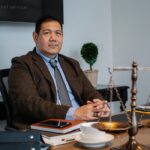 Middle aged ethnic legislator looking at camera and sitting at table with scales of justice in office